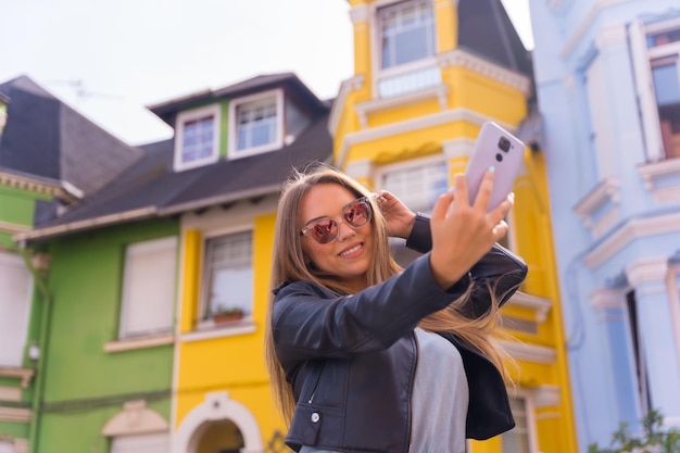 Joven mujer rubia con chaqueta de cuero tomando un selfie detrás de una fachada colorida