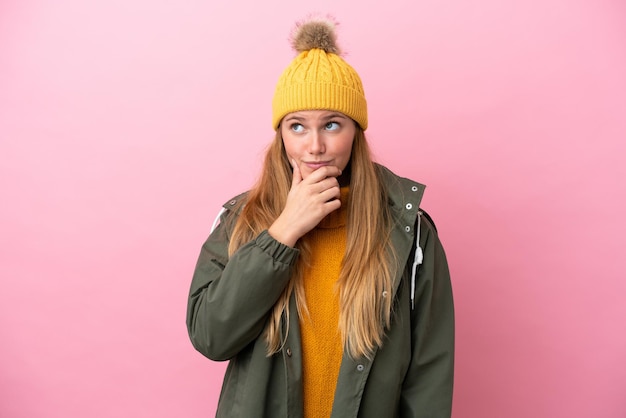 Joven mujer rubia con chaqueta de invierno aislada de fondo rosa que tiene dudas y con expresión facial confusa