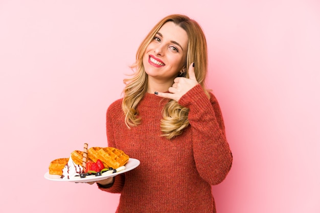 Joven mujer rubia comiendo un postre de gofres aislado mostrando un gesto de llamada de teléfono móvil con los dedos.