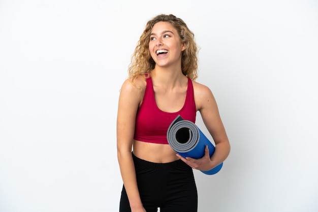 Joven mujer rubia de deporte que va a clases de yoga mientras sostiene una estera aislada sobre fondo blanco mirando hacia un lado y sonriendo