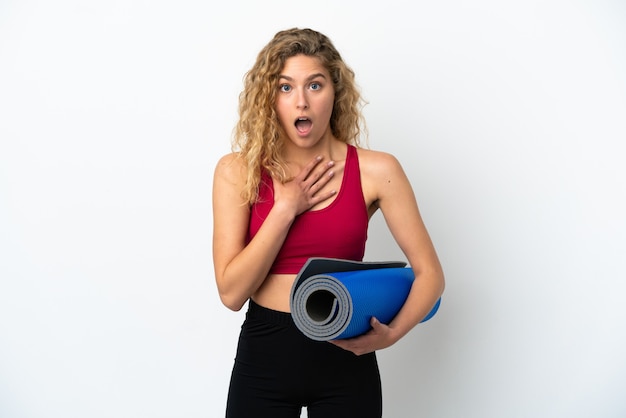 Joven mujer rubia de deporte que va a clases de yoga mientras sostiene una estera aislada sobre fondo blanco sorprendida y conmocionada mientras mira a la derecha