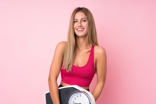 Joven mujer rubia deporte sobre pared rosa aislado con máquina de pesaje