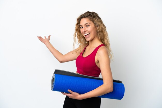 Joven mujer rubia deportiva que va a clases de yoga mientras sostiene una estera aislada sobre fondo blanco extendiendo las manos hacia un lado para invitar a venir