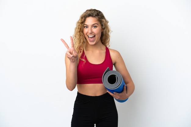 Joven mujer rubia deportiva que va a clases de yoga mientras sostiene una estera aislada sobre fondo blanco sonriendo y mostrando el signo de la victoria