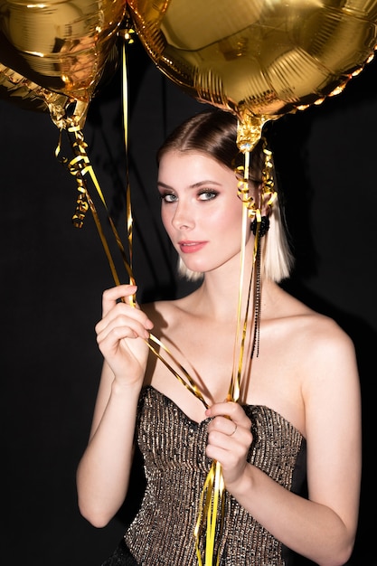 Joven mujer rubia elegante con globos de color dorado disfrutando de la fiesta de cumpleaños u otro evento de celebración sobre fondo negro