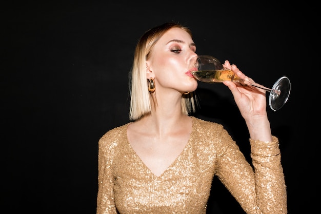 Joven mujer rubia en elegante vestido bebiendo champán después de brindar en la fiesta en la discoteca donde se celebra algún evento de la vida