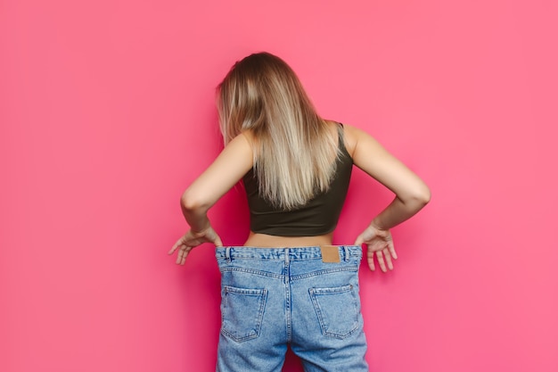 Una joven mujer rubia esbelta caucásica vestida con pantalones vaqueros anchos está de espaldas y demuestra una pérdida de peso exitosa aislada en una pared de color rosa brillante