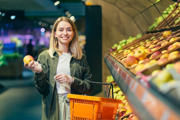 Joven mujer rubia escoge elige frutas y verduras en el mostrador del supermercado. Ama de casa femenina de compras en el mercado de pie cerca de los grandes almacenes vegetales con una canasta en las manos. Examina la manzana