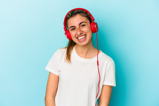 Joven mujer rubia escuchando música en auriculares aislados en la pared azul feliz, sonriente y alegre