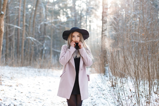 Joven mujer rubia con estilo en un elegante sombrero negro con un elegante abrigo rosa con un vestido negro de moda está de pie en el bosque de invierno. Hermosa chica disfruta del paisaje.