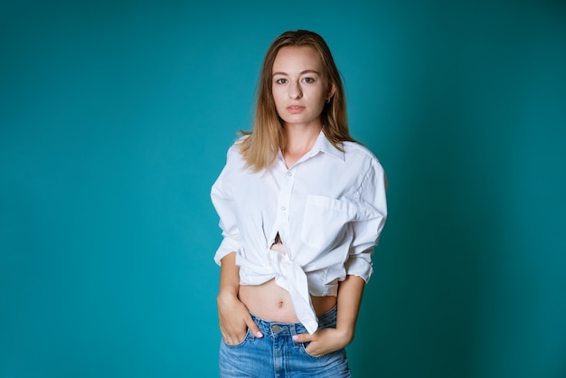 Joven mujer rubia de etnia caucásica posando con una camisa blanca y jeans en una pared azul