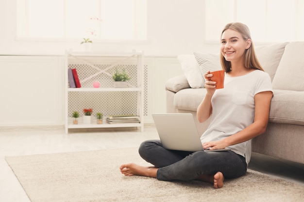 Joven mujer rubia feliz trabajando en una computadora portátil sentada en el piso navegando por la red y tomando espacio para copiar café