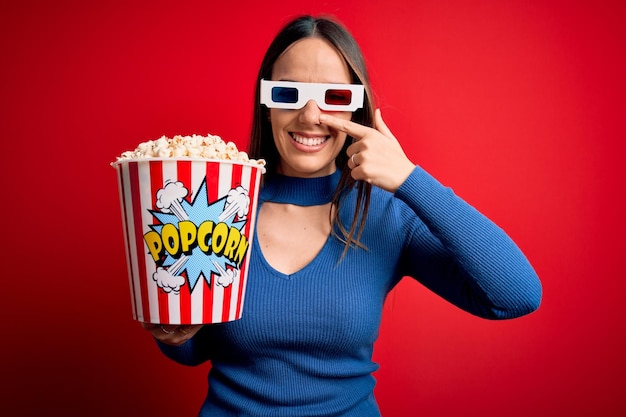 Joven mujer rubia con gafas 3d y comiendo un paquete de palomitas de maíz viendo una película en el cine Señalando con el dedo de la mano a la cara y la nariz sonriendo alegre Concepto de belleza