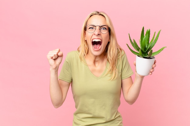 Joven mujer rubia gritando agresivamente con una expresión de enojo y sosteniendo un cactus