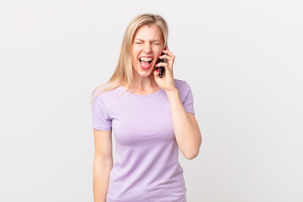 Joven mujer rubia gritando agresivamente, luciendo muy enojada y llamando con un teléfono inteligente.