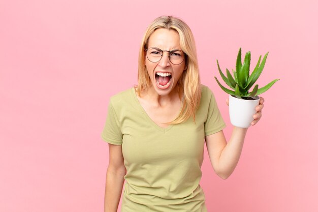 Joven mujer rubia gritando agresivamente, luciendo muy enojada y sosteniendo un cactus