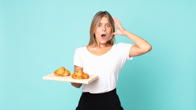 Joven mujer rubia gritando con las manos en el aire y sosteniendo una bandeja de croissant