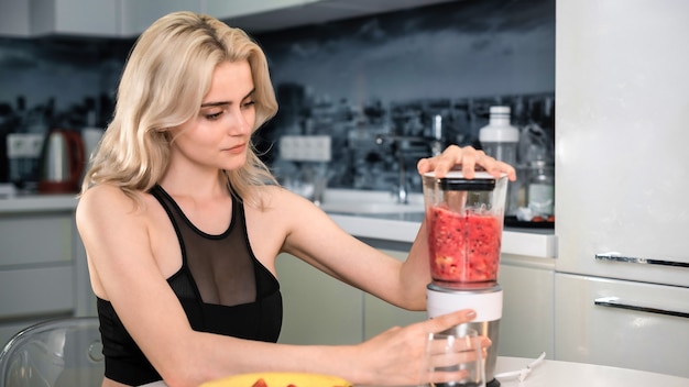 Una joven mujer rubia haciendo un batido de frutas y bayas, arándano, fresa, plátano