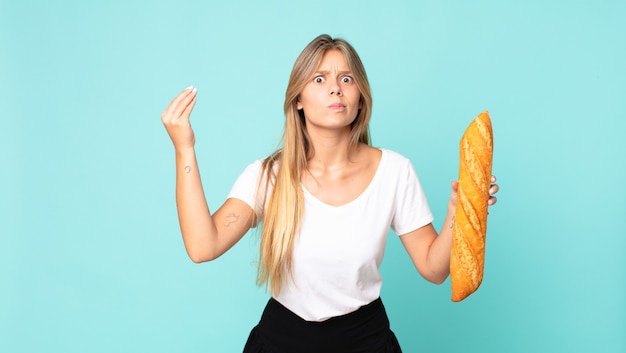 Joven mujer rubia haciendo gesto de capice o dinero, diciéndole que pague y sosteniendo una baguette de pan