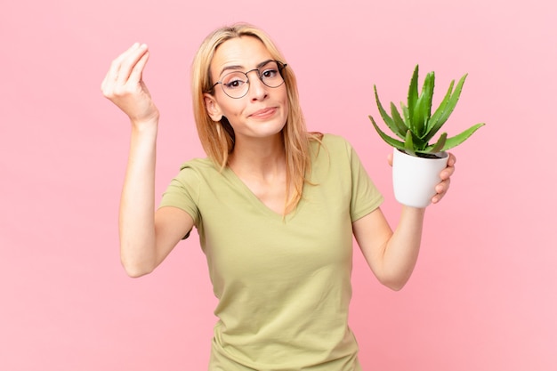 Joven mujer rubia haciendo gesto de capice o dinero, diciéndole que pague y sosteniendo un cactus