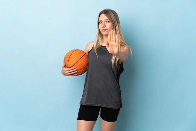 Joven mujer rubia jugando baloncesto aislado en azul haciendo gesto de parada con la mano