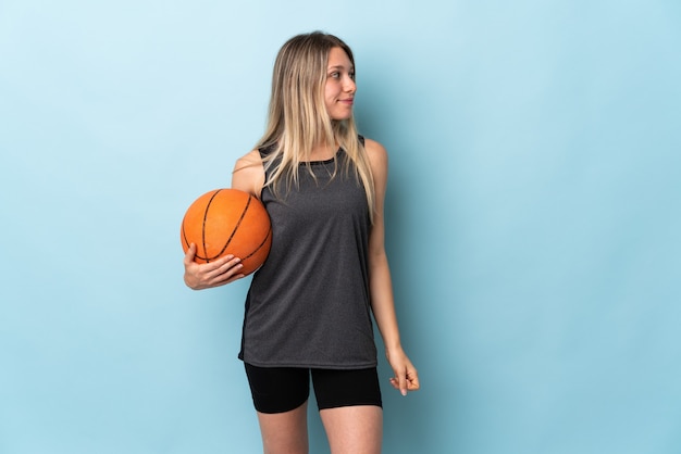 Joven mujer rubia jugando baloncesto aislado en la pared azul mirando hacia el lado