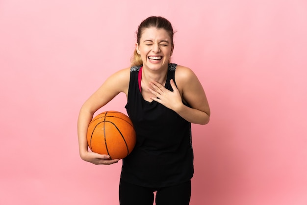 Joven mujer rubia jugando baloncesto sobre fondo rosa sonriendo mucho