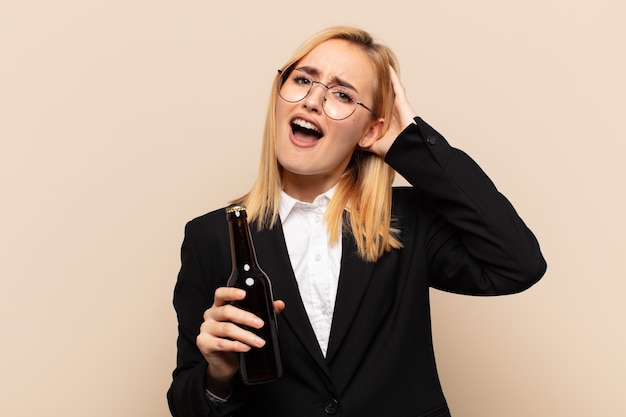 Joven mujer rubia levantando las manos a la cabeza, con la boca abierta, sintiéndose extremadamente afortunada, sorprendida, emocionada y feliz