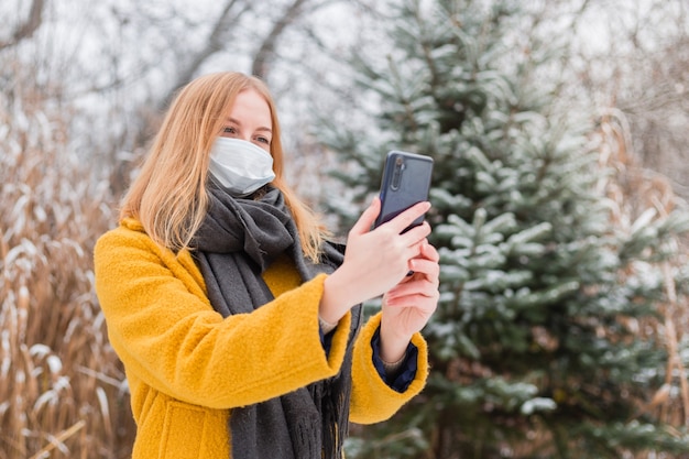 Joven mujer rubia con mascarilla médica blanca con aplicación de teléfono inteligente sobre la naturaleza