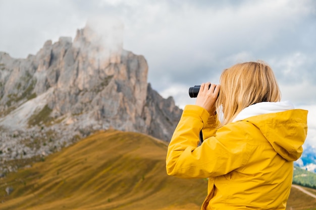 Joven mujer rubia mira con binoculares al paso de alta montaña en los Dolomitas, el paso de Giau