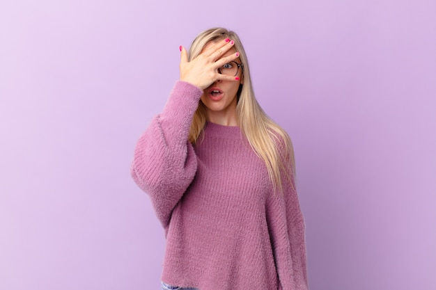 Joven mujer rubia mirando conmocionado, asustado o aterrorizado, cubriéndose la cara con la mano