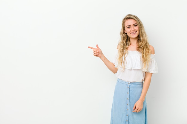 Joven mujer rubia mirando emocionado y sorprendido apuntando hacia un lado y hacia arriba para copiar espacio contra la pared blanca