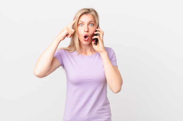 Joven mujer rubia mirando sorprendida, dándose cuenta de un nuevo pensamiento, idea o concepto y llamando con un teléfono inteligente