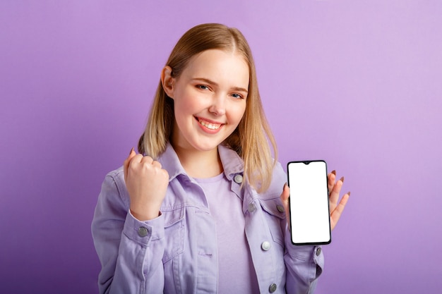 Joven mujer rubia muestra maqueta de pantalla de teléfono inteligente vacía sobre fondo de color púrpura aislado.