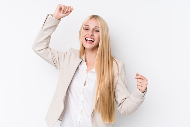 Joven mujer rubia de negocios en la pared blanca celebrando un día especial, salta y levanta los brazos con energía.