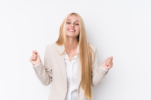 Joven mujer rubia de negocios en la pared blanca levantando el puño, sentirse feliz y exitoso. Concepto de victoria