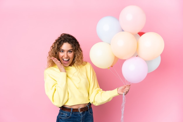 Joven mujer rubia con pelo rizado atrapando muchos globos aislados en rosa con expresión facial sorpresa