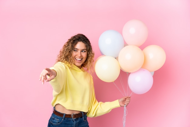 Joven mujer rubia con el pelo rizado atrapar muchos globos en el dedo señalador rosa a un lado