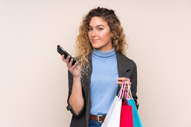 Joven mujer rubia con el pelo rizado en color beige con bolsas de compras y escribir un mensaje con su teléfono celular a un amigo