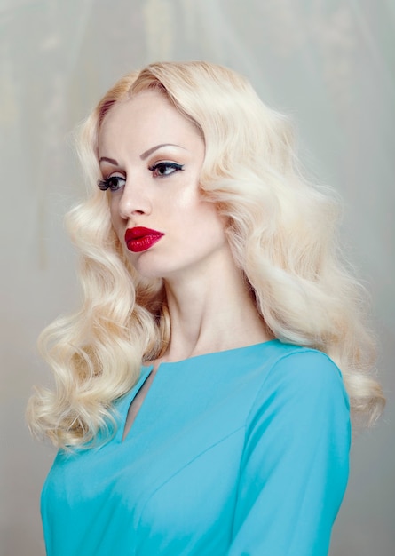Joven mujer rubia con pelo rizado y maquillaje, labios rojos, vestida con un elegante vestido azul.