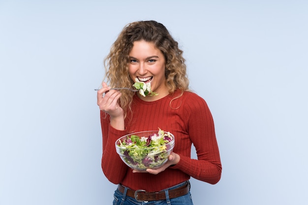 Joven mujer rubia con pelo rizado sosteniendo una ensalada sobre pared aislada