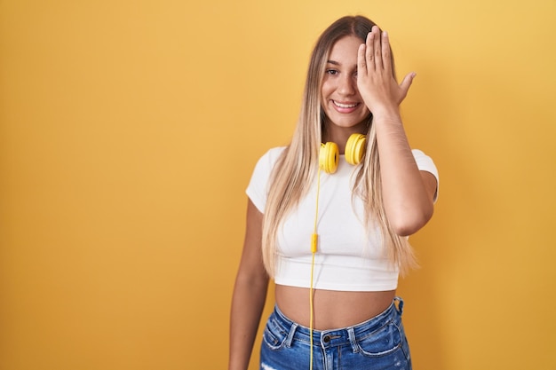 Joven mujer rubia de pie sobre fondo amarillo con auriculares que cubren un ojo con la mano, sonrisa segura en la cara y emoción sorpresa.