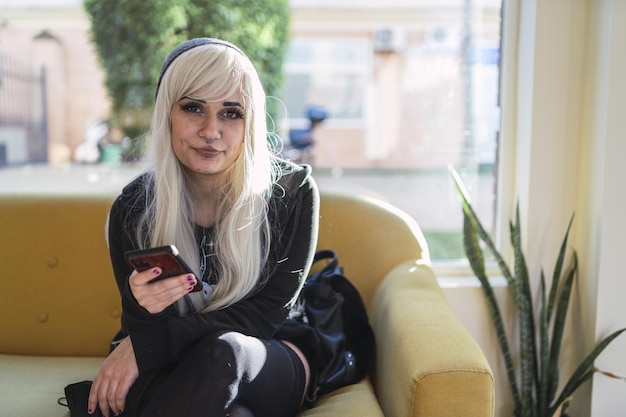 Joven mujer rubia con piercings sentada en un café y desplazándose por su teléfono