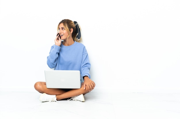 Joven mujer rubia con el portátil aislado manteniendo una conversación con el teléfono móvil