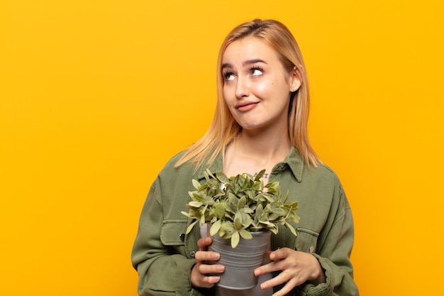 Joven mujer rubia preguntándose, pensando en pensamientos e ideas felices, soñando despierto, buscando copiar espacio en el lateral