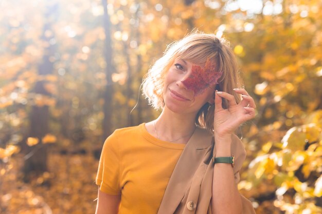 Joven mujer rubia que cubre un ojo con otoño de hoja de arce roja y concepto de temporada caída al aire libre