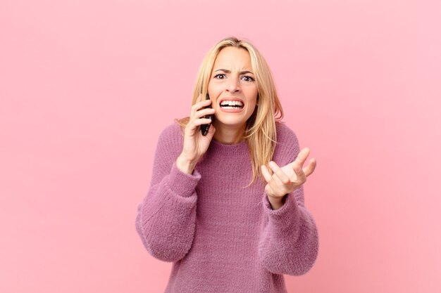 Joven mujer rubia que parece desesperada, frustrada y estresada y hablando con un teléfono inteligente