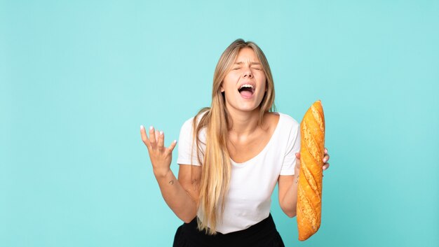 Joven mujer rubia que parece desesperada, frustrada y estresada y sosteniendo una baguette de pan