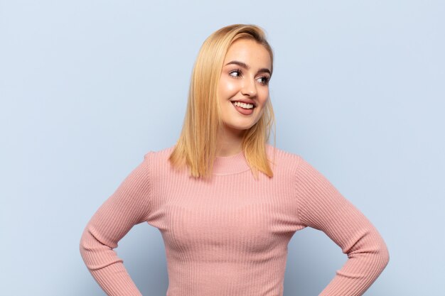 Joven mujer rubia que parece feliz, alegre y segura, sonriendo con orgullo y mirando al lado con ambas manos en las caderas