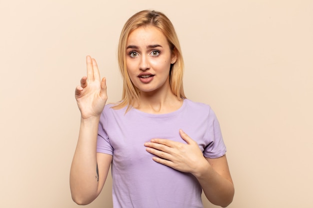 Joven mujer rubia que parece feliz, segura y digna de confianza, sonriendo y mostrando el signo de la victoria, con una actitud positiva